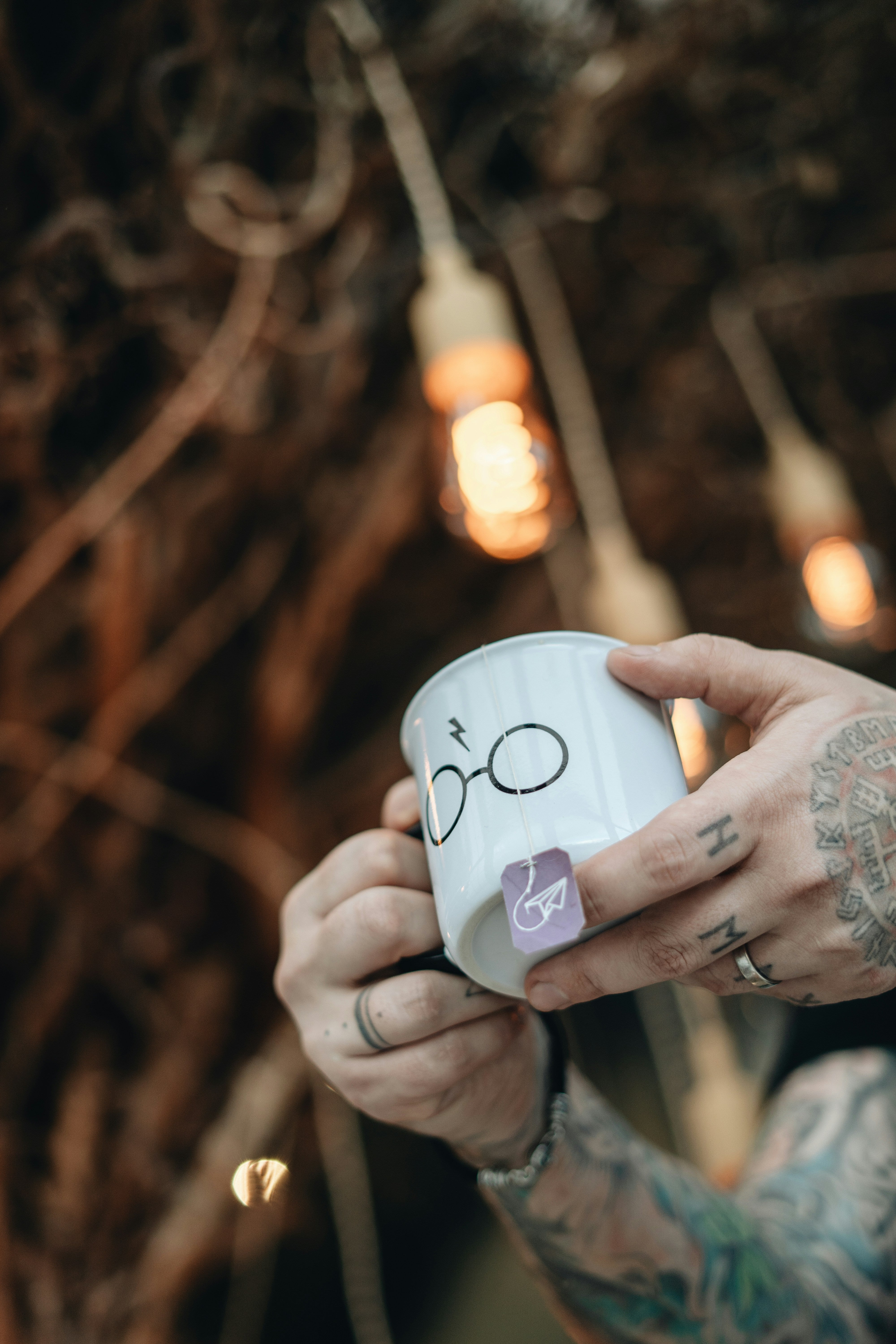 Tattooed person holding a mug of coffee and shilajit outdoors beneath warm hanging lights
